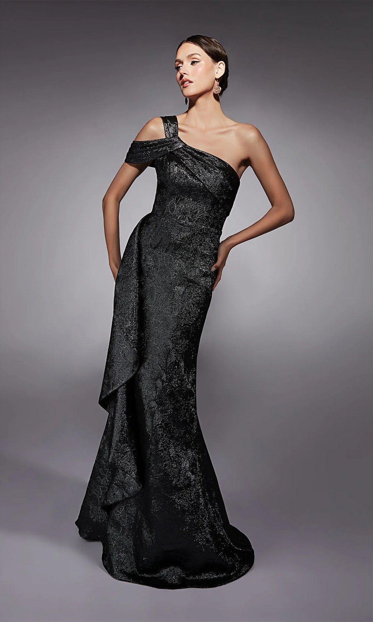 A woman poses against a plain gray background wearing the Alyce 27734—a black, textured, one-shoulder long formal dress with an asymmetrical design.